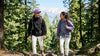 Man and woman wearing windcheater jackets hiking in pine forest with snowy mountains in India