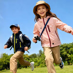 Girl wearing pink Venzina waterproof hooded windbreaker and boy in navy jacket running outdoors under blue sky and green trees
