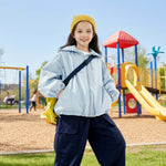 Girl wearing a light blue kids outdoor jacket with hood, standing on a playground — breathable windbreaker jacket for school and travel.