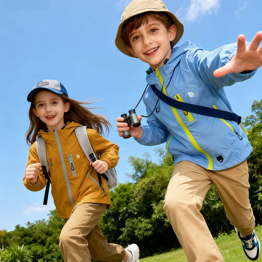 Two kids wearing Venzina waterproof hooded windbreaker jackets, one blue with yellow zip and one brown, running outdoors under blue sky
